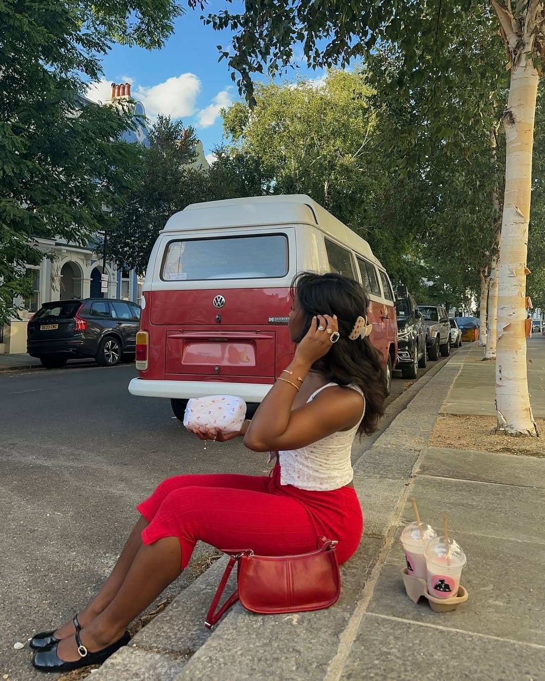 Pessoa sentada na calçada, com bolsa vermelho tomate e van vintage ao fundo.