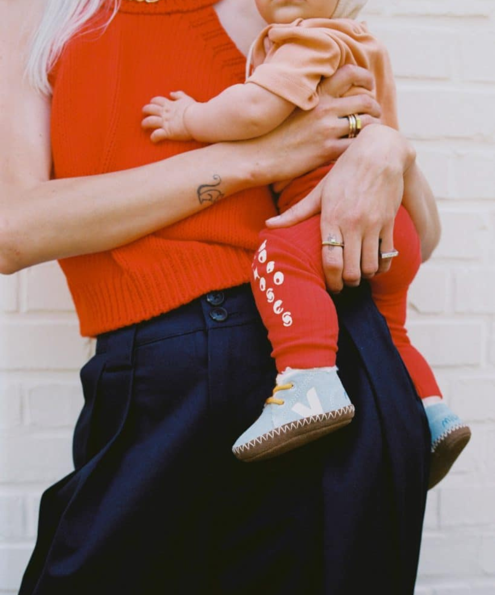 Criança no colo de um adulto, vestindo calça vermelha e tênis lúdicos azuis com detalhes brancos.