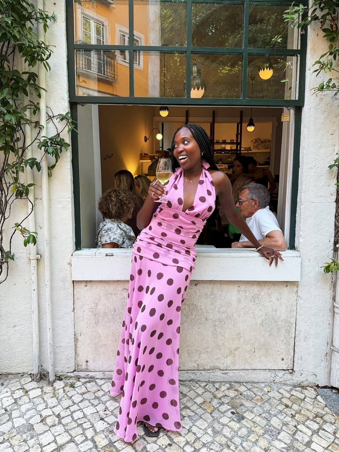 Mulher sorrindo e segurando uma taça de vinho, usando um vestido de poá rosa em frente a uma janela de restaurante.