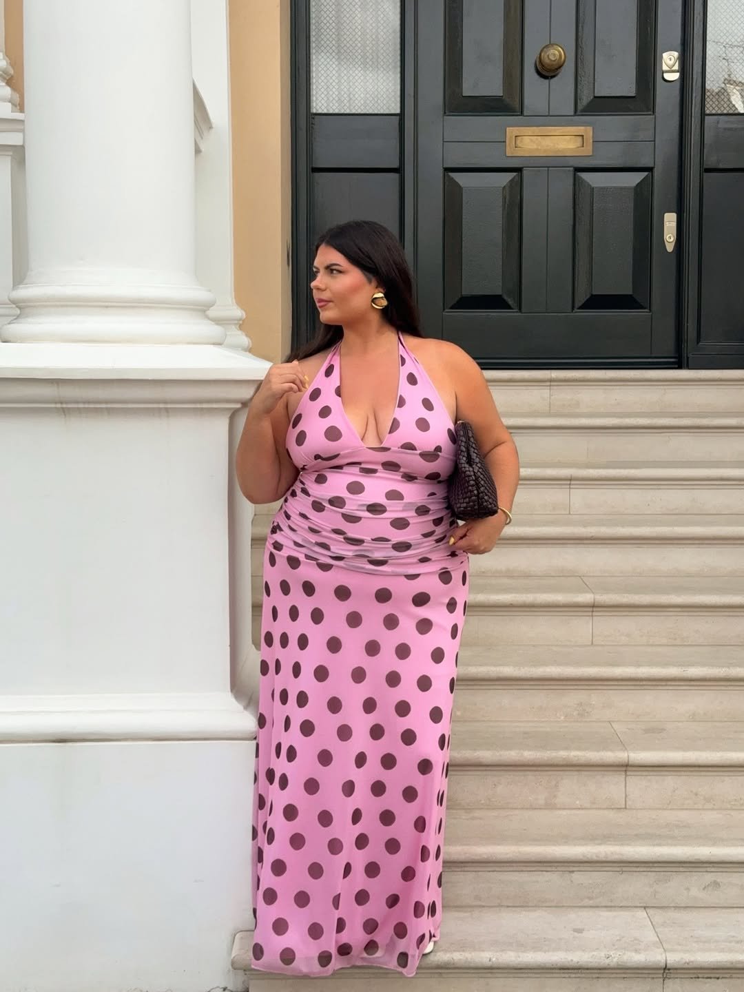 Mulher usando vestido de poá rosa com bolinhas escuras diante de uma porta preta elegante.