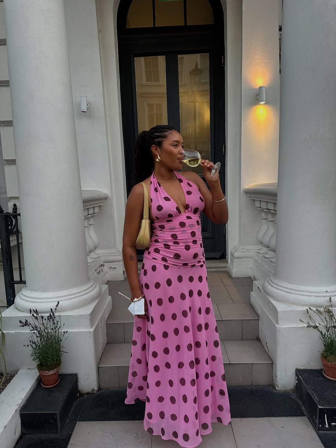 Mulher em frente a coluna branca vestindo vestido de poá rosa, tomando vinho.