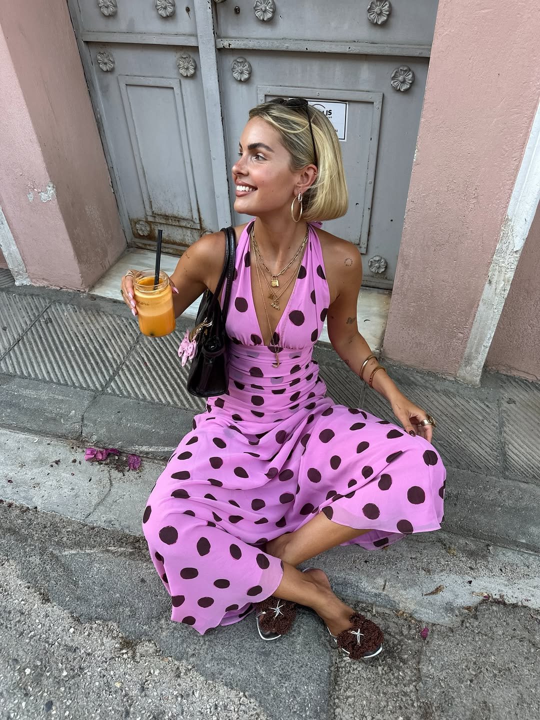 Pessoa usa vestido de poá rosa com bolas pretas, segurando uma bebida laranja, sorrindo sentada na calçada.