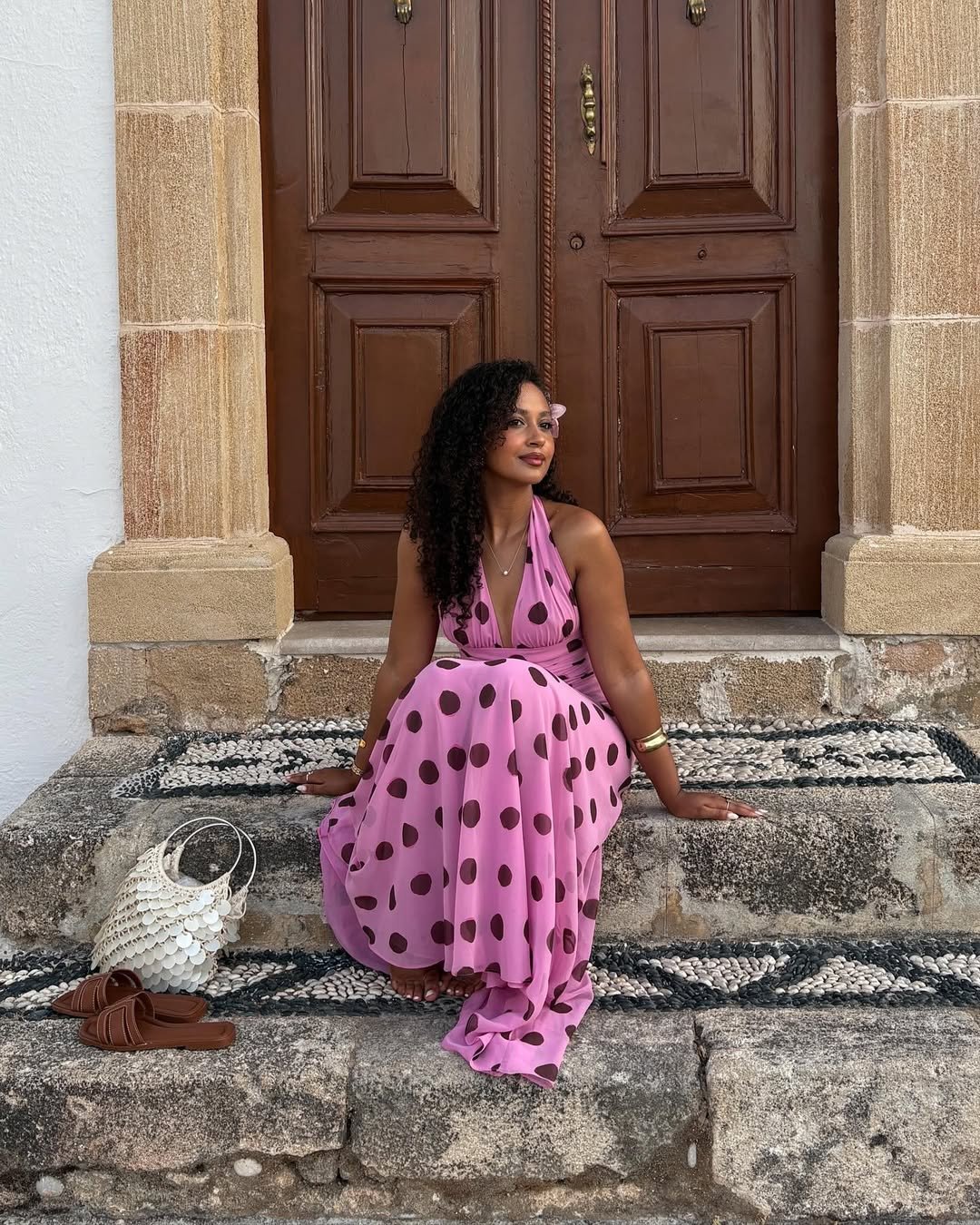 Mulher sentada em degraus de pedra, usando vestido de poá rosa e marrom, com uma bolsa clara ao lado.