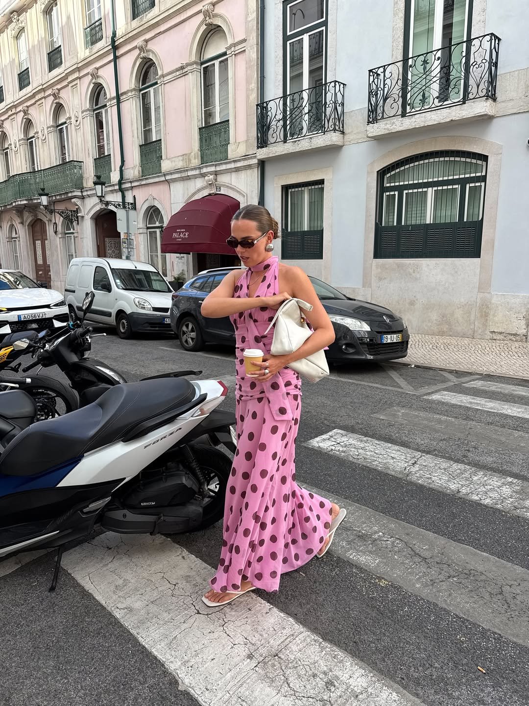 Pessoa usando vestido de poá rosa, óculos escuros e chinelos, segura bebida e bolsa branca em rua com moto e carros.