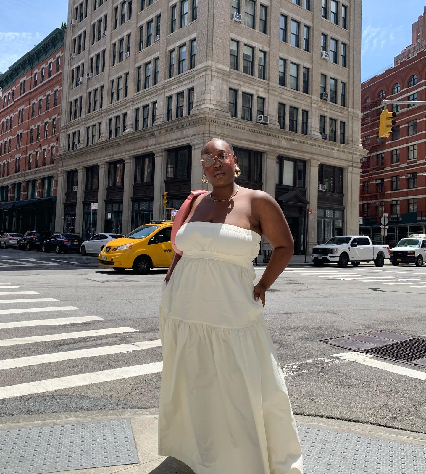 Pessoa em um vestido sem alça branco na cidade, com prédios e táxi ao fundo, em um dia ensolarado.