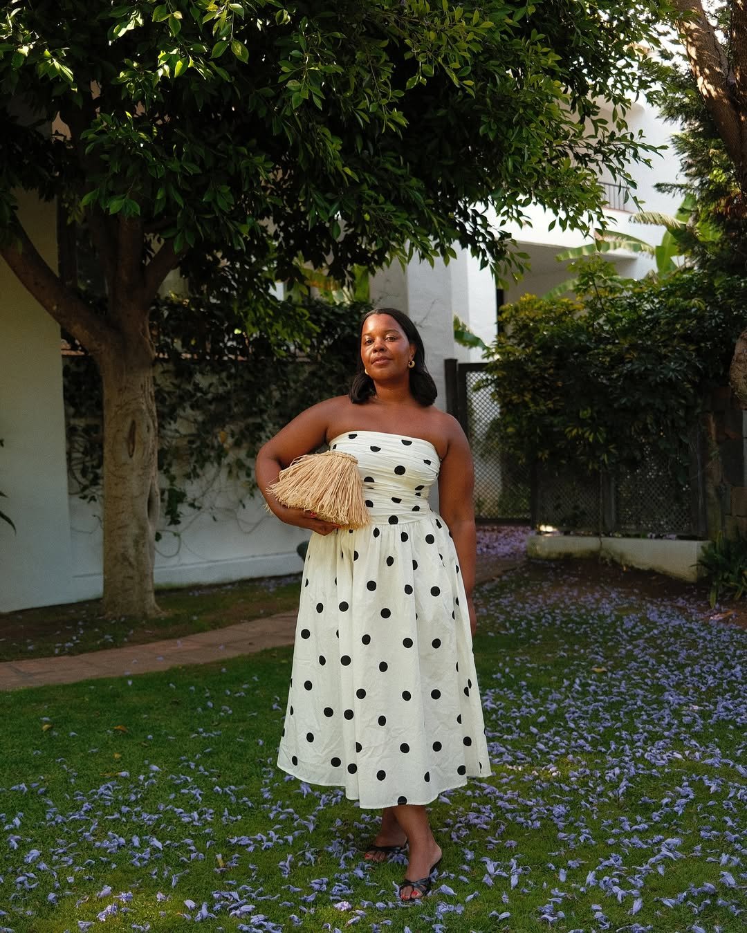 Pessoa usando vestido sem alça de bolinhas em um jardim florido, segurando uma bolsa de palha.