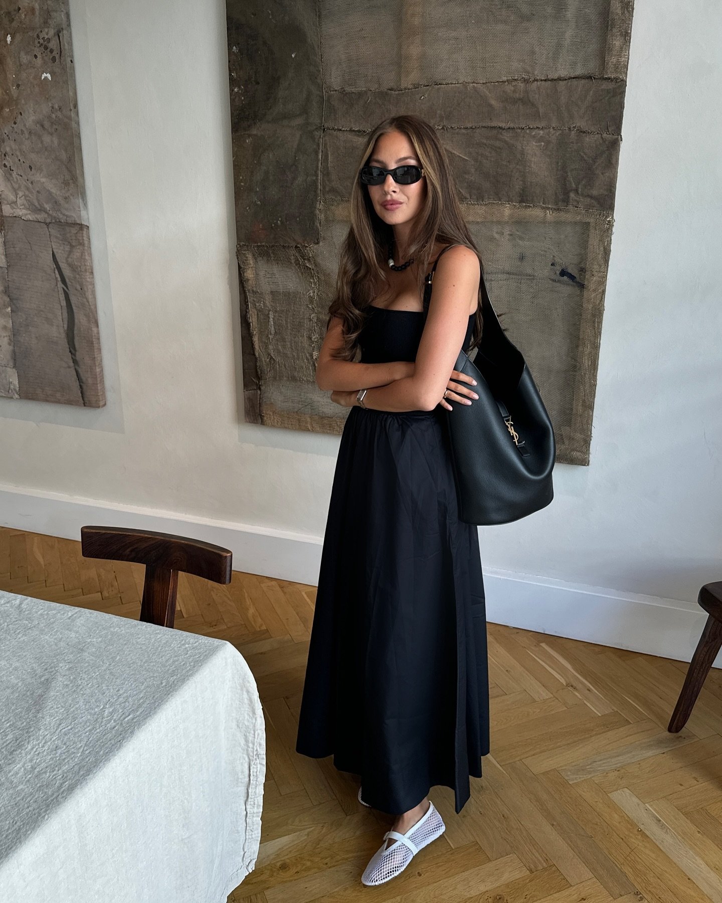 Pessoa usando um vestido preto sem alça, óculos de sol e bolsa preta, em um ambiente com arte abstrata.