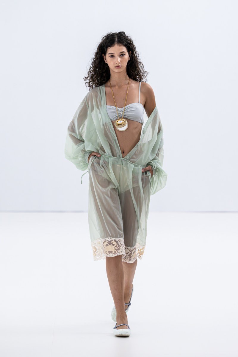 Modelo com vestido translúcido azul-claro e colar dourado durante a Milan Fashion Week SS26.