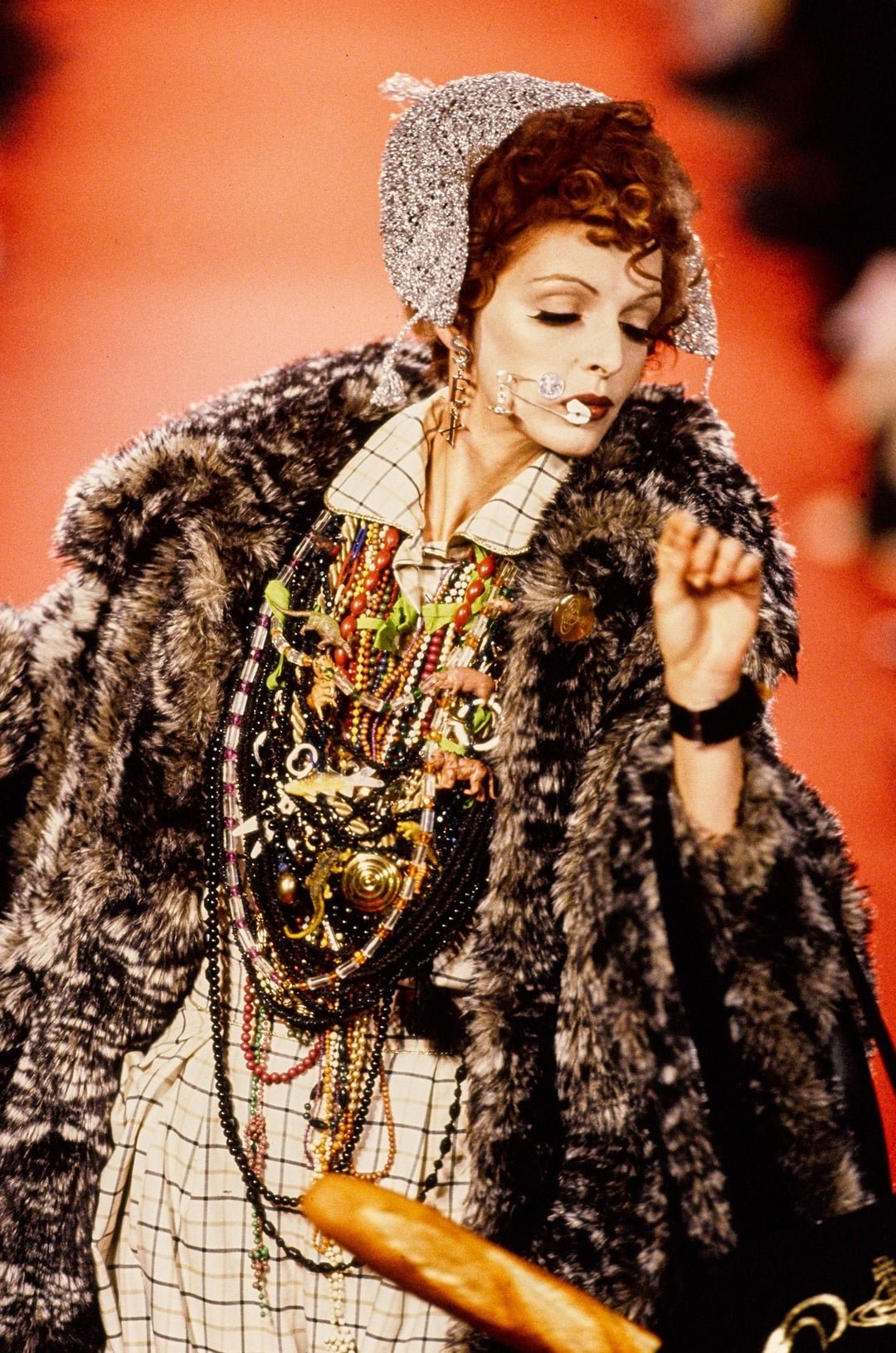 Desfile de moda apresenta modelo com visual exuberante, usando casaco de pele falsa e vários piercings faciais estilizados.