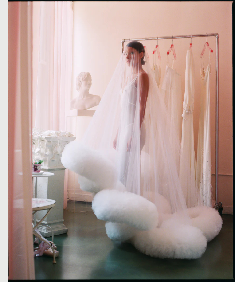 Pessoa veste um longo véu branco sob vestido não tradicional com detalhes volumosos, em um quarto decorado.