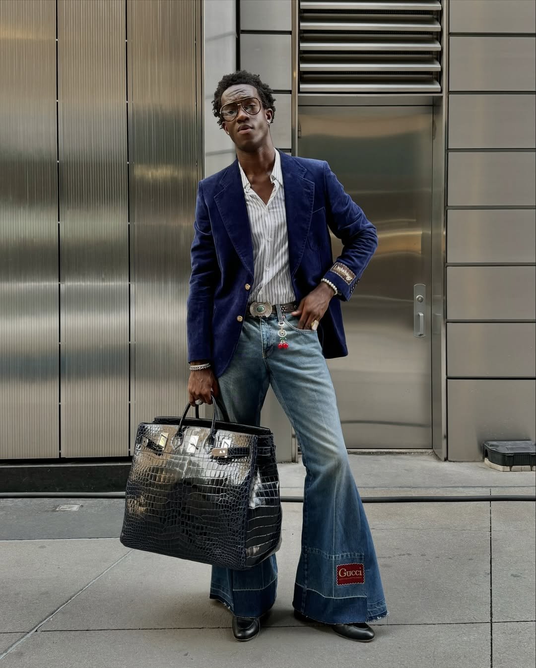 Wisdom Kaye vestindo um blazer azul, jeans largos, carregando uma grande bolsa preta.
