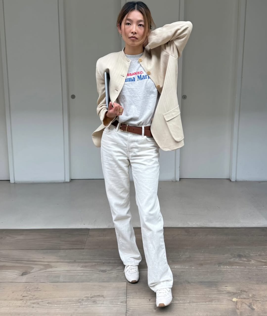 Pessoa vestindo blazer bege sobre graphic tee, calça branca e tênis, segura um notebook em ambiente interno.