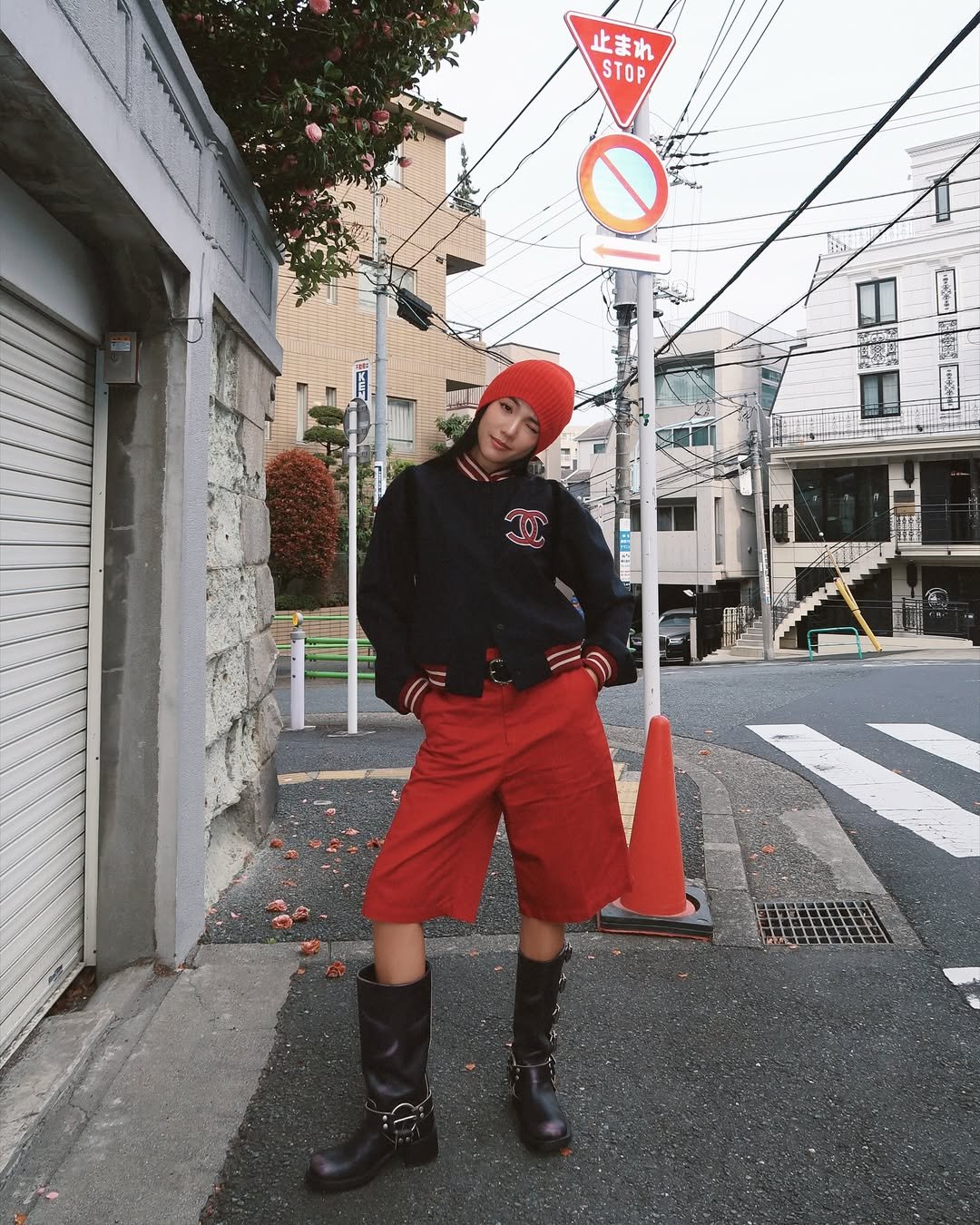 Pessoa em look estiloso com gorro e bermuda vermelhos, moletom preto e botas. Ambiente urbano ao fundo.