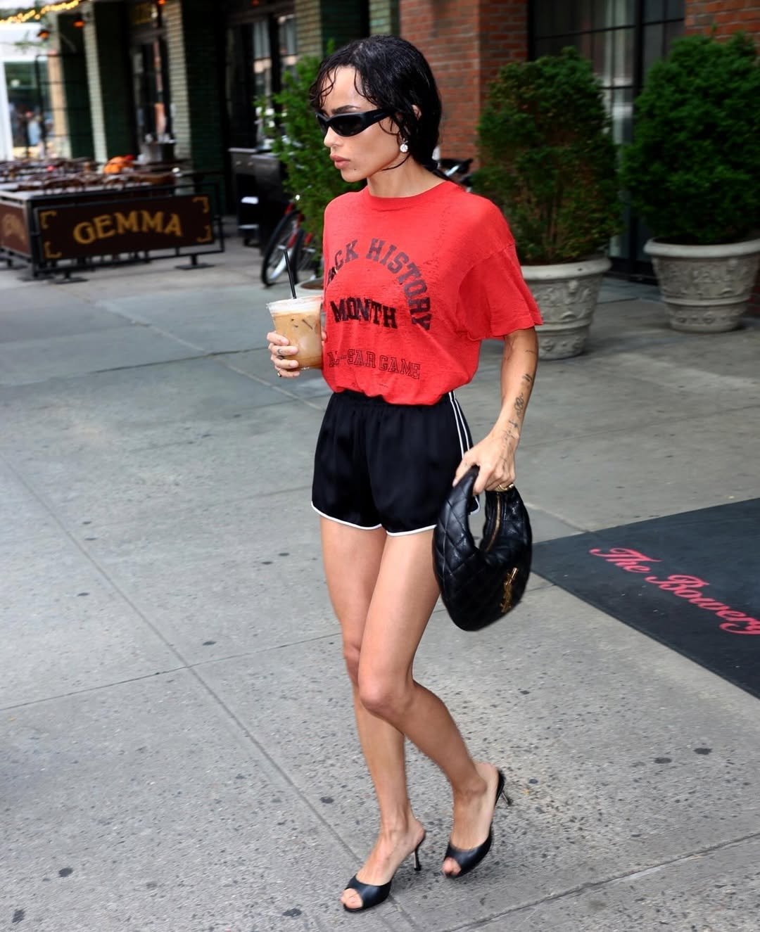 Pessoa usando graphic tee vermelha, short preto e óculos escuros, caminhando na calçada enquanto segura bebida gelada.