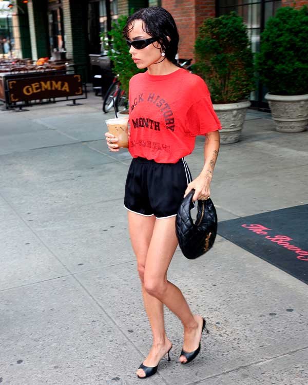 Mulher com óculos escuros, blusa vermelha e shorts pretos, segura bebida e bolsa. Caminha em rua urbana.