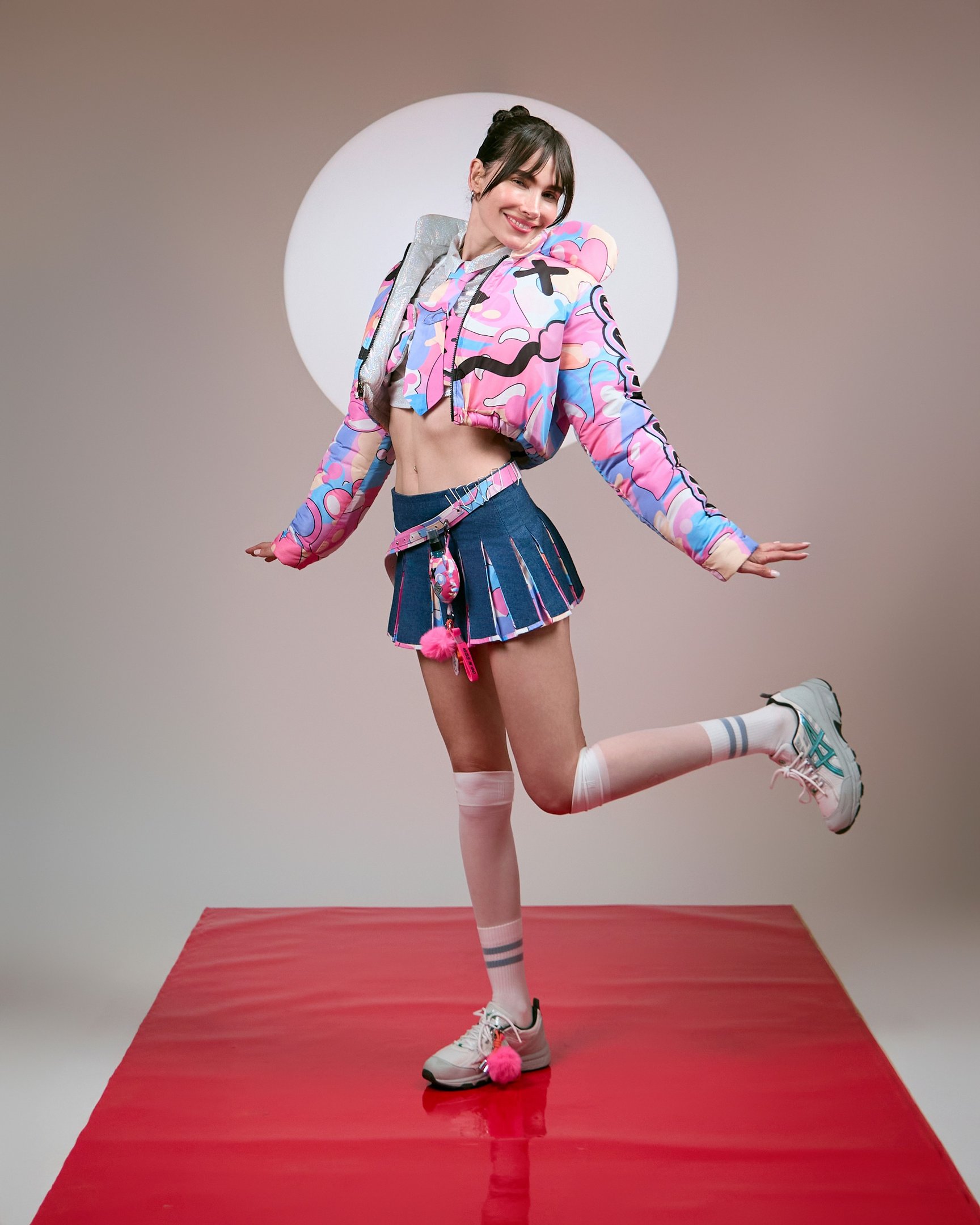 A chaveiro estiloso é um acessório que complementa looks vibrantes e modernos. Na imagem, a modelo exibe um visual ousado com uma jaqueta cropped em tons de rosa, combinada com uma saia de denim. Os detalhes, como os pompons e meias longas, criam um ar jo