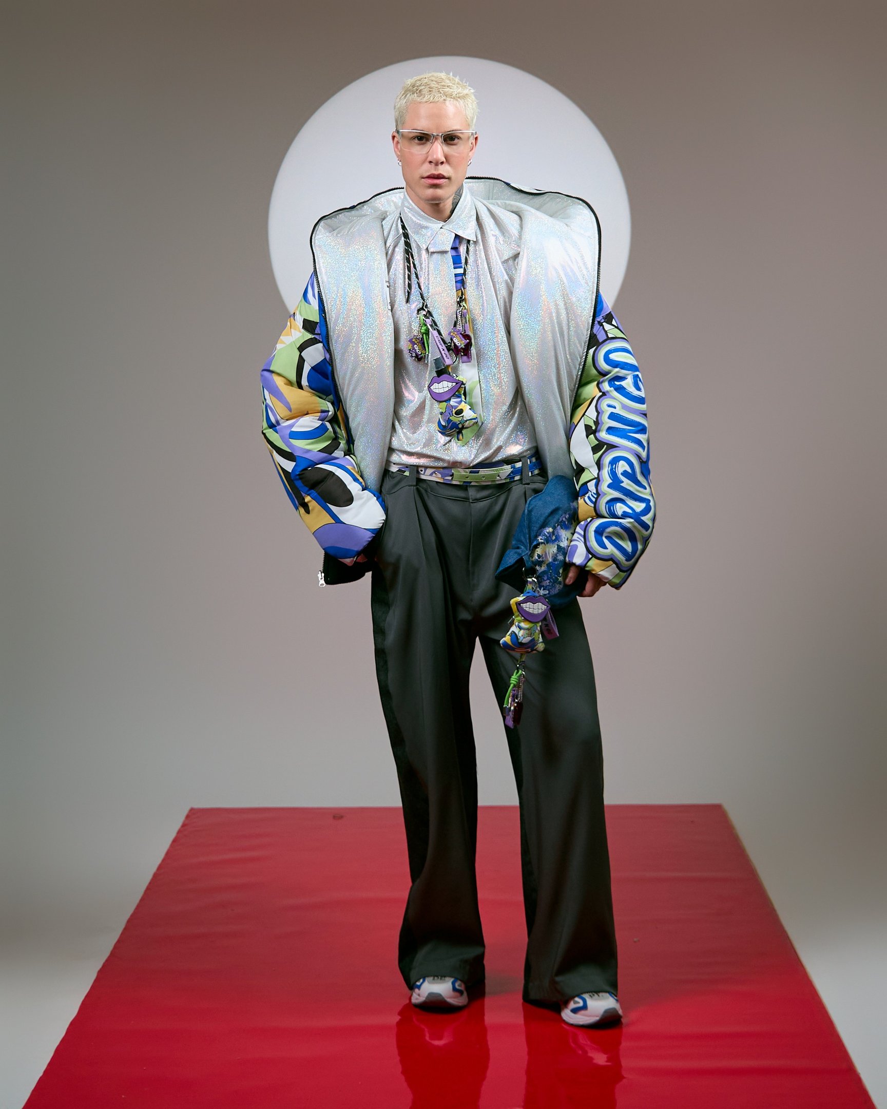 Um chaveiro estiloso é destacado na moda contemporânea, pendurado em um colar que complementa um look ousado. A peça central é uma jaqueta brilhante, com estampas vibrantes, combinada com uma camisa metálica e calças largas. O modelo, com cabelo platinado