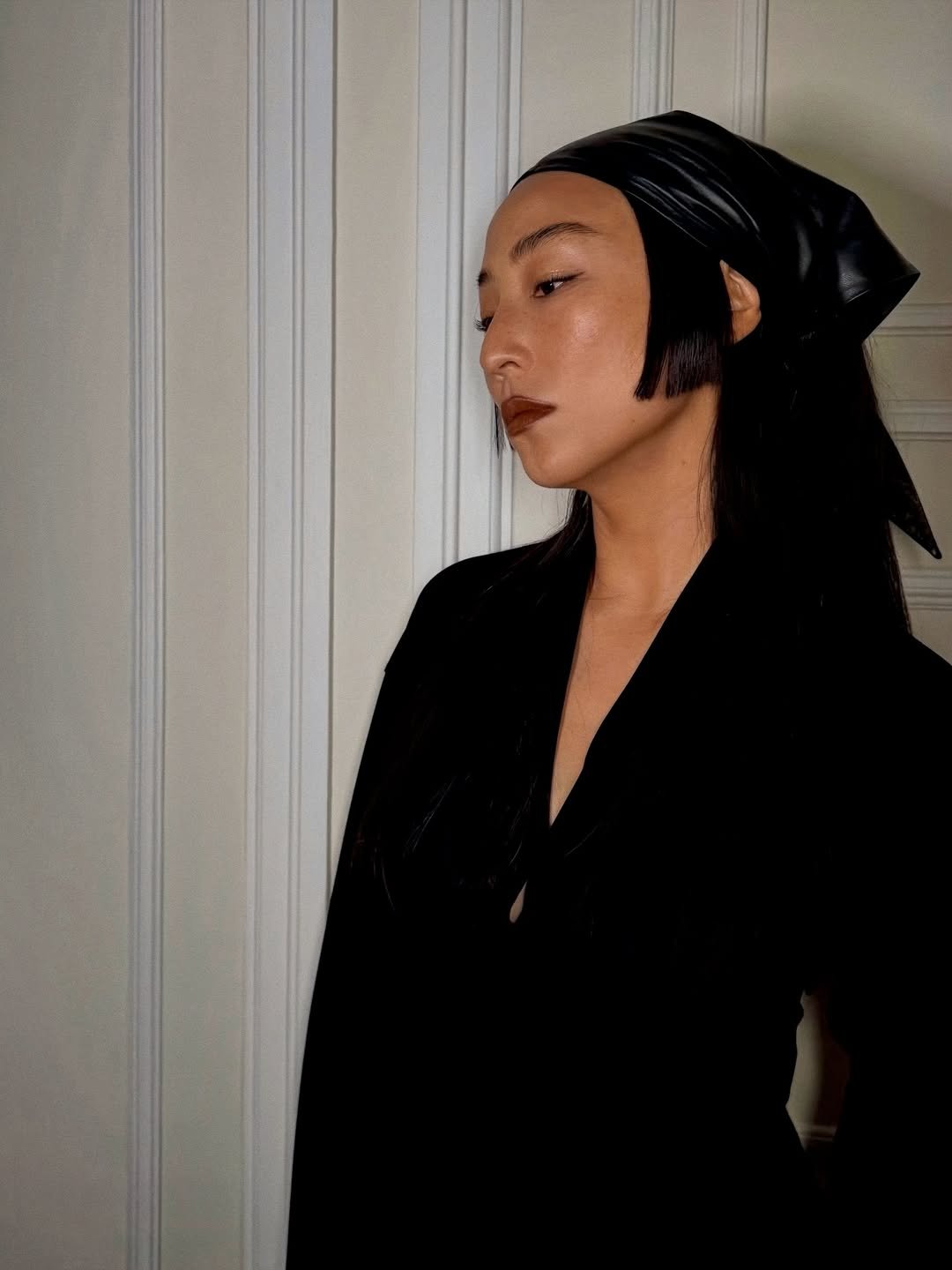 O estilo da Greta Lee é marcado por uma estética minimalista e sofisticada. Na imagem, ela usa um vestido preto que destaca sua silhueta, complementado por um lenço de couro preto na cabeça, que confere um toque contemporâneo e ousado. A maquiagem é sutil