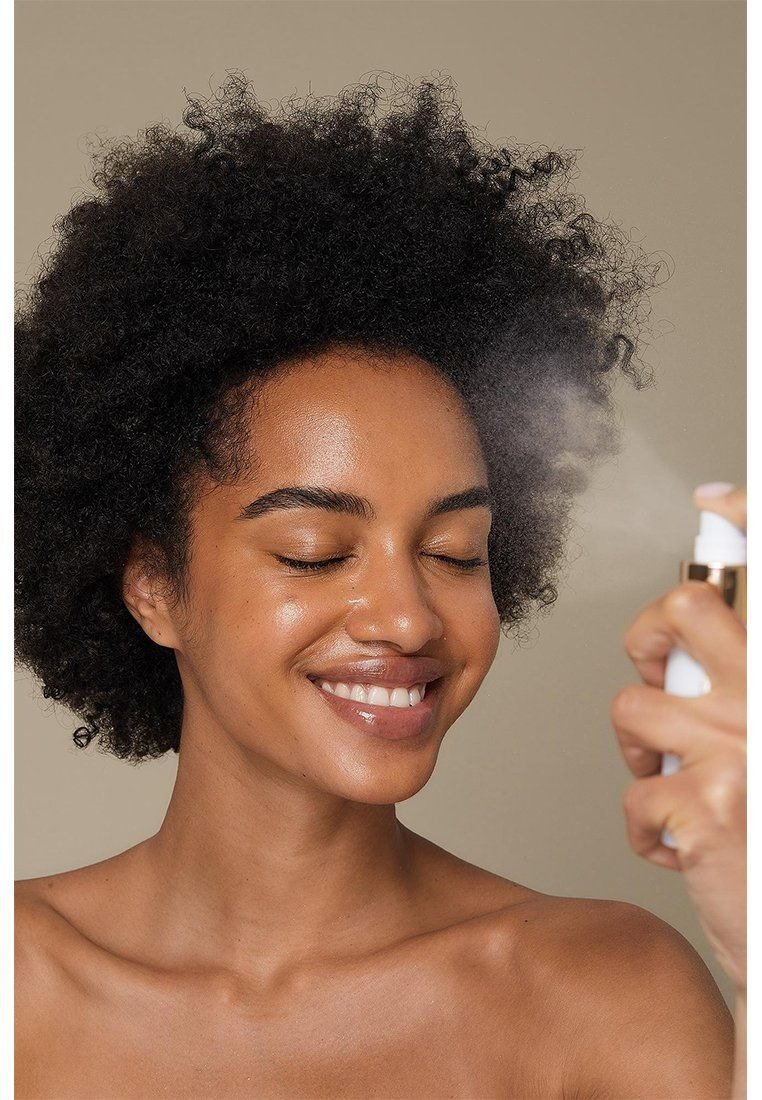 A imagem retrata uma mulher sorridente aplicando água termal em seu rosto com um borrifador. Sua pele, levemente brilhante e bem cuidada, destaca-se em um fundo neutro.