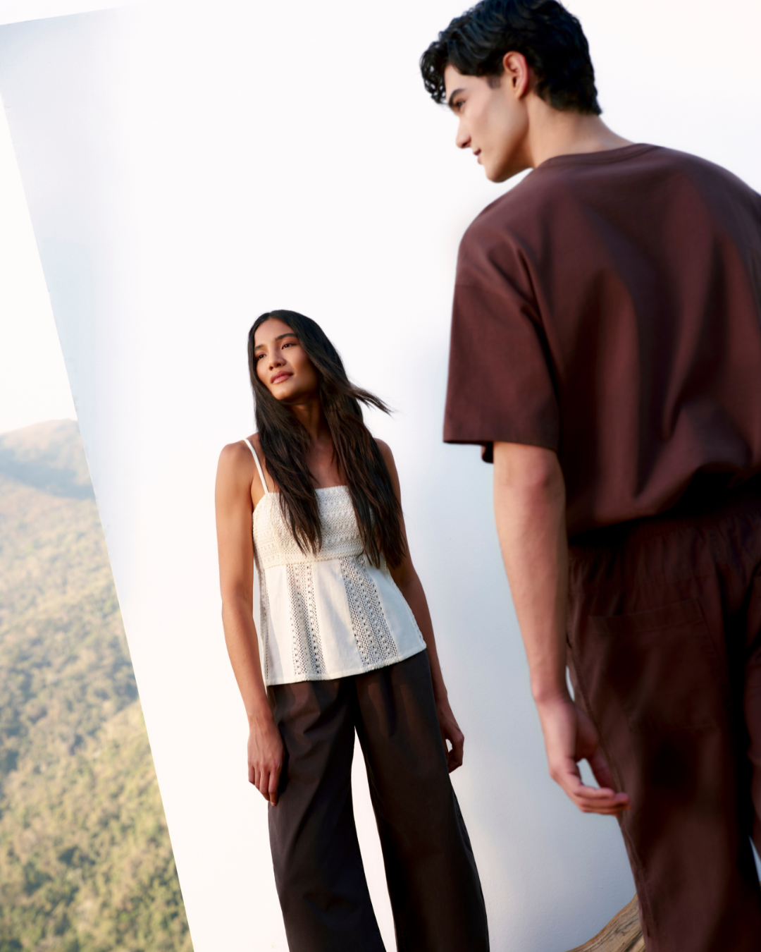 Um casal posa em um cenário natural, destacando a moda sustentável com peças de algodão regenerativo. A mulher usa uma blusa clara com detalhes rendados e calças escuras, enquanto o homem veste uma camiseta marrom e calças combinando. A luz suave ressalta