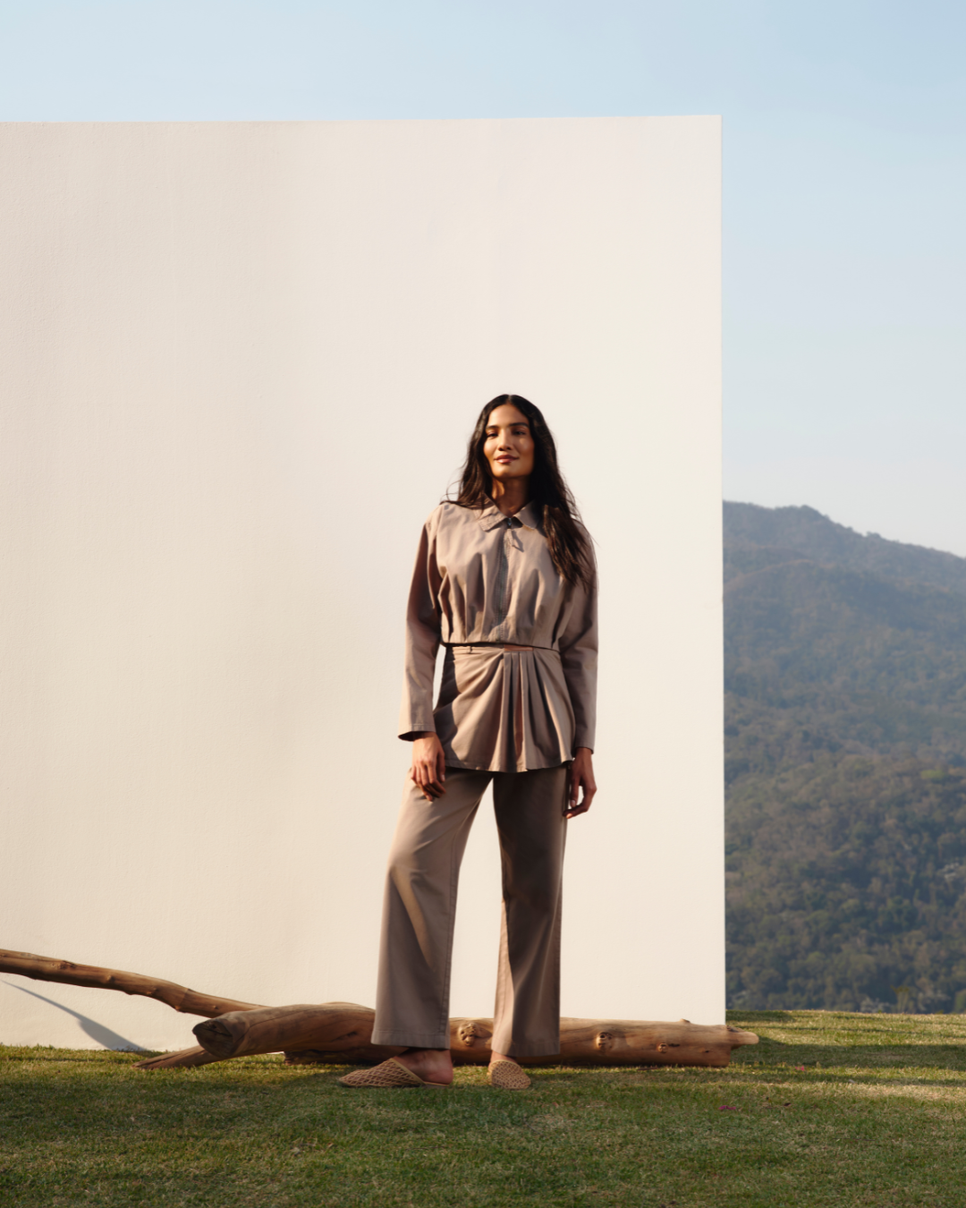 A imagem apresenta uma modelo em um cenário natural, vestindo um conjunto de roupas em tons terrosos, feito de algodão regenerativo. A blusa, com detalhes plissados, e as calças largas ressaltam a elegância e o conforto, refletindo a tendência sustentável