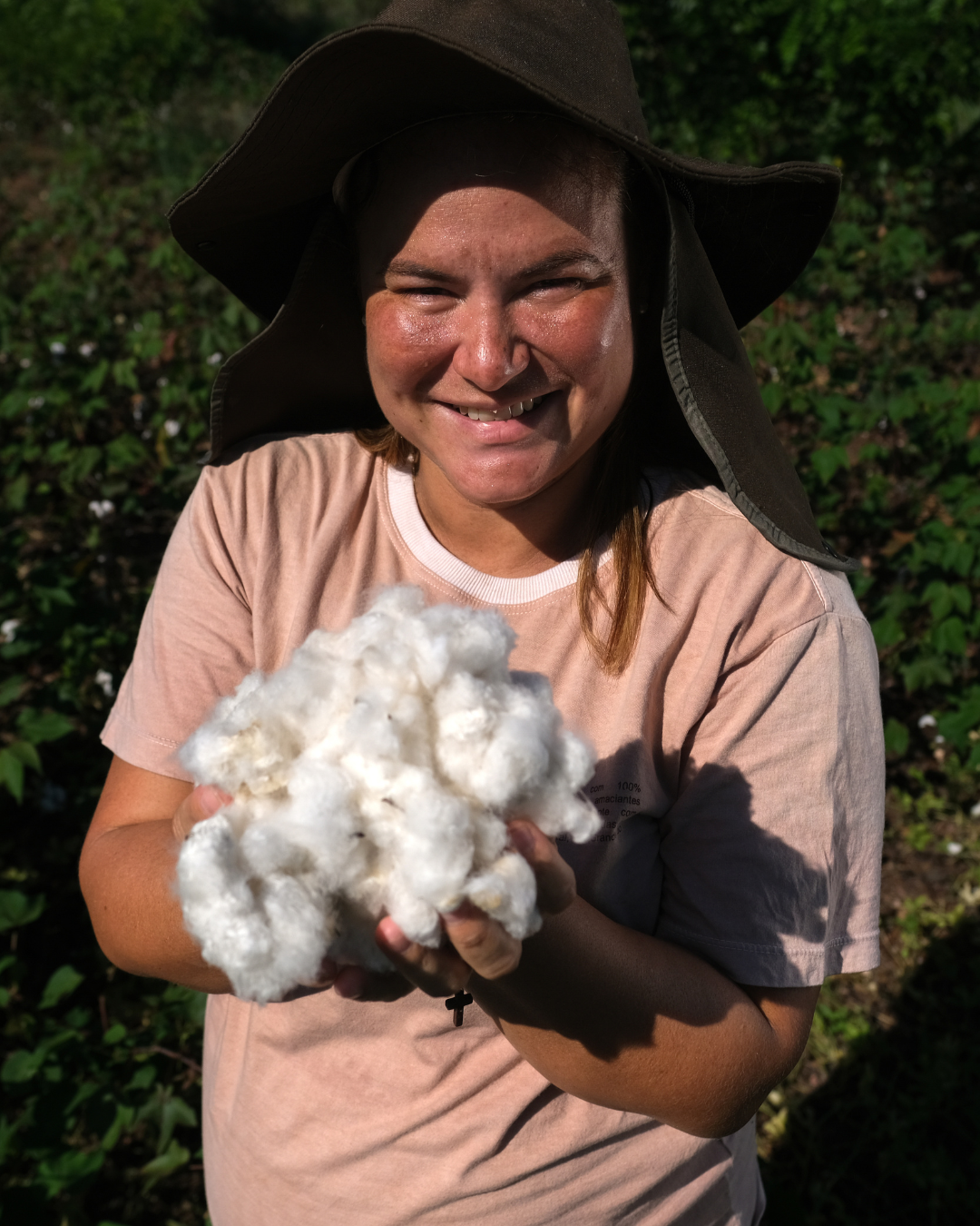 Uma mulher sorridente segura um punhado de algodão regenerativo, destacando a maciez e a textura do material. Ela está em um campo verdejante, com um chapéu de sol que a protege. O algodão regenerativo é essencial na moda sustentável