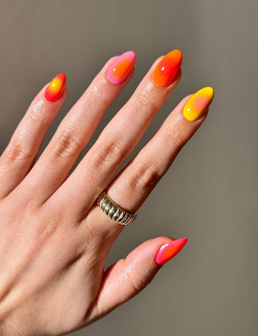As unhas aura nails exibem um design vibrante com um gradiente que transita entre tons quentes de rosa, laranja e amarelo. A forma afunilada das unhas destaca a elegância do estilo, enquanto um anel dourado complementa a composição.