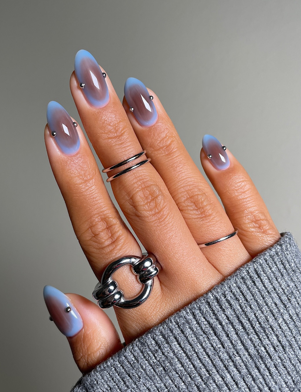 As unhas aura nails apresentam um efeito degradê em tons de azul e marrom, com formato alongado e elegante. Pequenos detalhes metálicos em forma de esferas adicionam um toque moderno. A mão, adornada com anéis prateados, complementa a estética de moda
