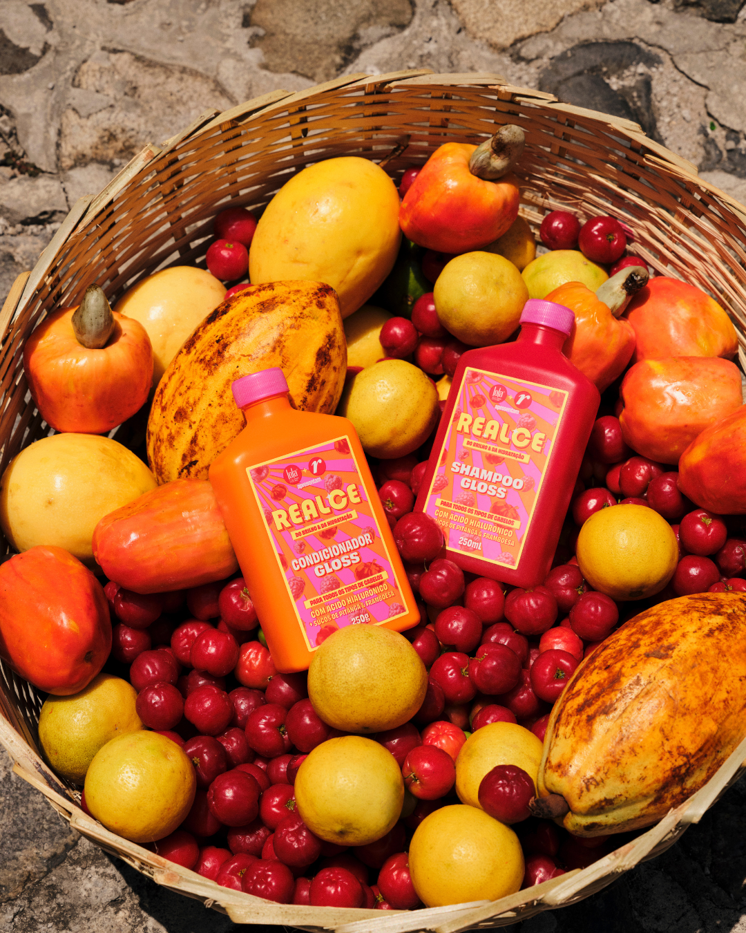 A imagem apresenta uma cesta de vime repleta de frutas tropicais vibrantes, como mangas, cajás e cerejas, ao lado de dois produtos de cabelo da linha Realce: um shampoo e um condicionador gloss, para dar banho de brilho no cabelo