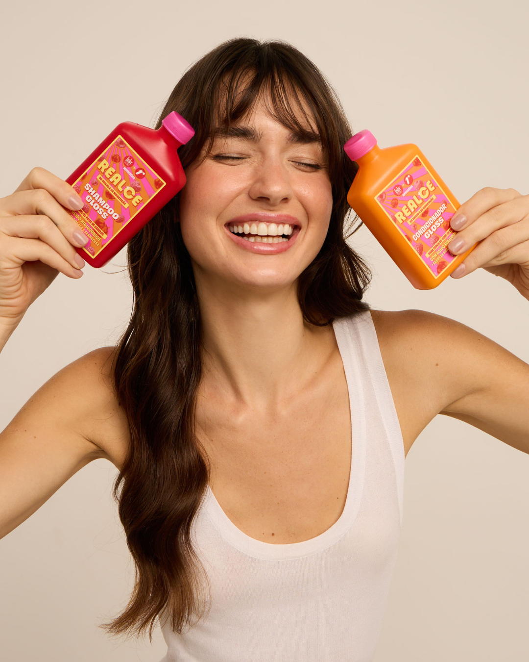 Uma mulher sorridente segura um frasco vermelho de shampoo e um frasco laranja de condicionador, ambos com rótulos vibrantes, para dar banho de brilho no cabelo