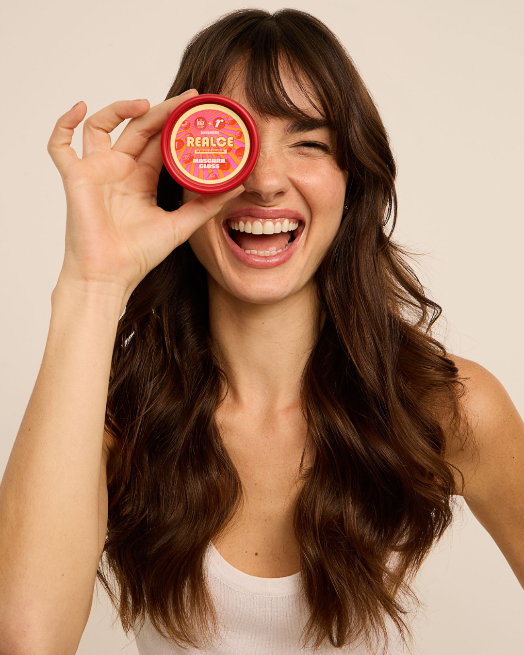 Uma mulher sorridente segura um pote de máscara de brilho para cabelo, destacando o produto, para dar banho de brilho no cabelo. Com cabelo castanho ondulado e bem cuidado, ela usa uma blusa branca, transmitindo uma imagem vibrante e positiva.