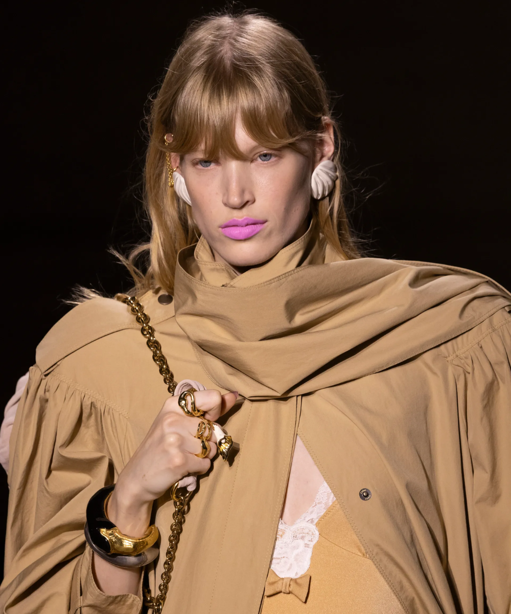 Desfile com modelo usando batom rosa vibrante, criticado nos anos 2010. Acessório dourado ao ombro complementa o visual.