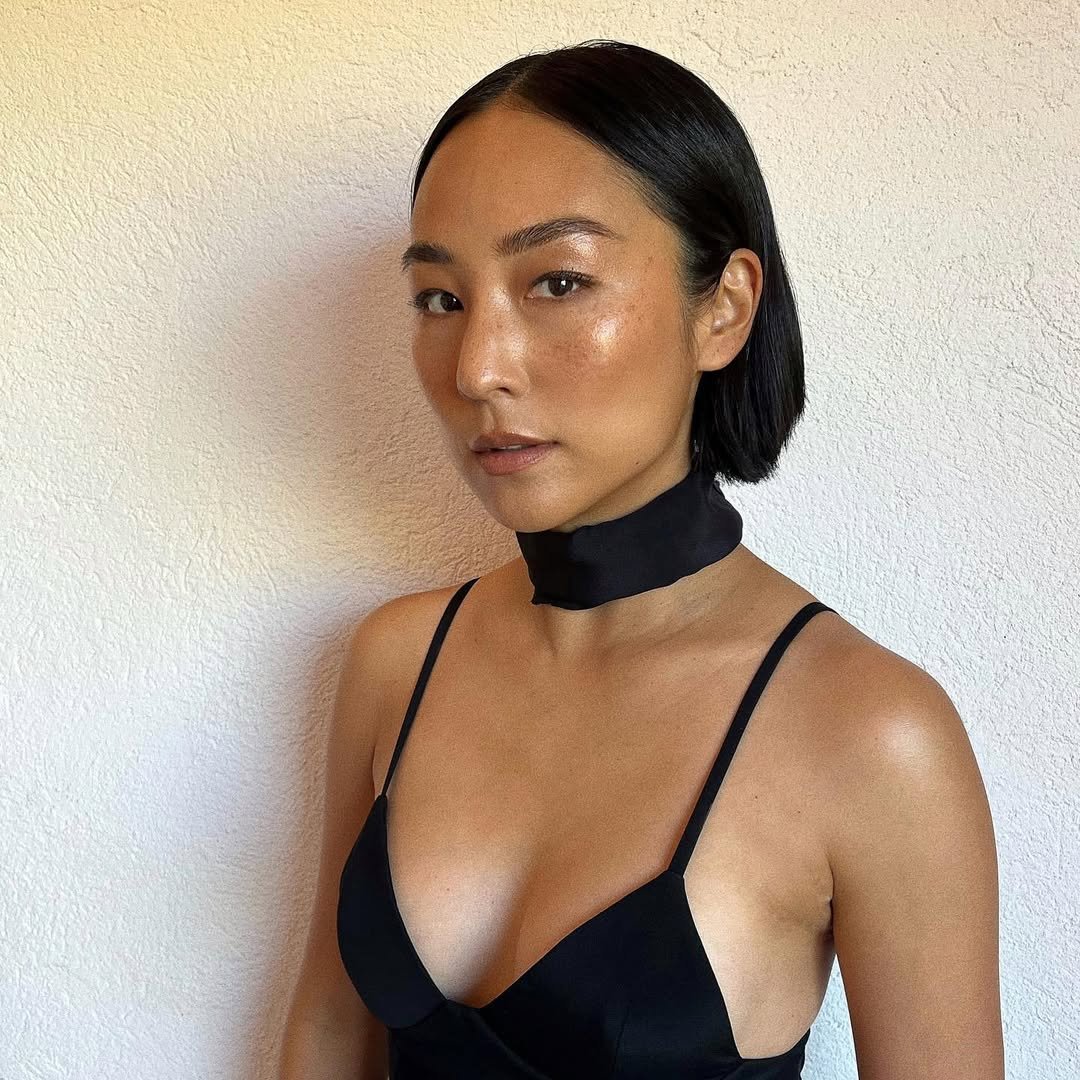 Greta Lee exibe um estilo sofisticado e minimalista, destacando-se com um vestido preto de alças finas que valoriza a silhueta. O uso de um choker preto adiciona um toque moderno ao look. Sua pele radiante e maquiagem leve ressaltam a beleza natural, refl