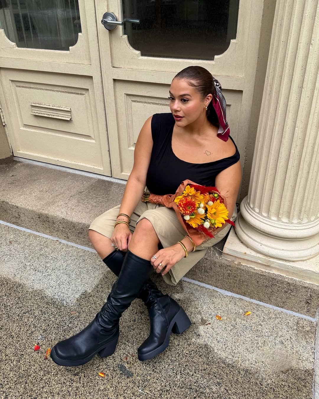 Pessoa sentada em escada, usando bermuda alfaiataria bege e blusa preta. Segura buquê de flores amarelas.