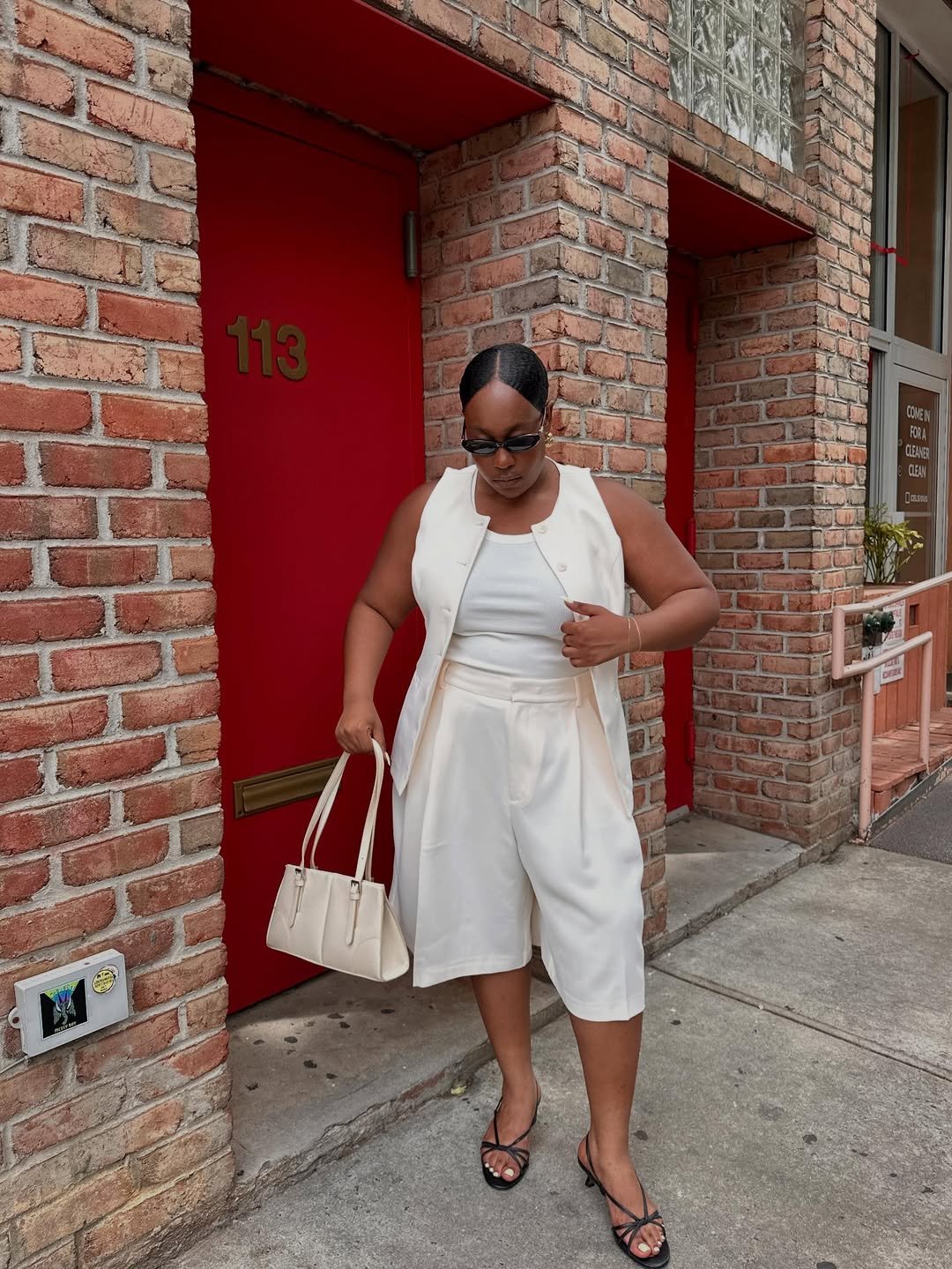 Pessoa usando bermuda alfaiataria branca e blusa combinando, ao lado de porta vermelha. Carrega bolsa creme.