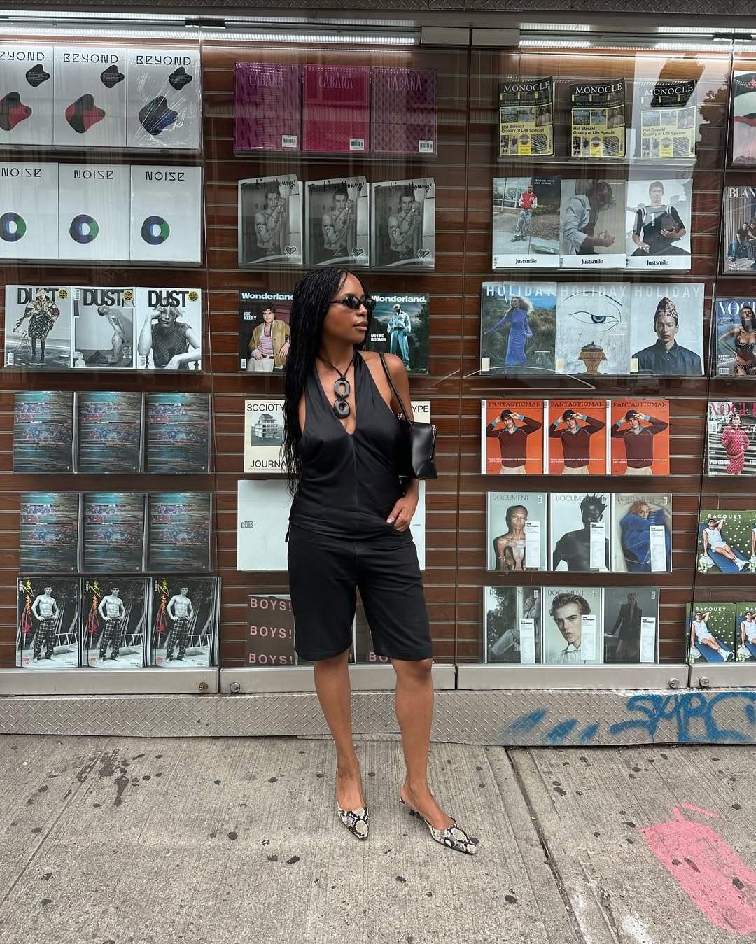 Mulher com óculos de sol e colar preto posa em frente a banca com revistas, usando bermuda alfaiataria preta.