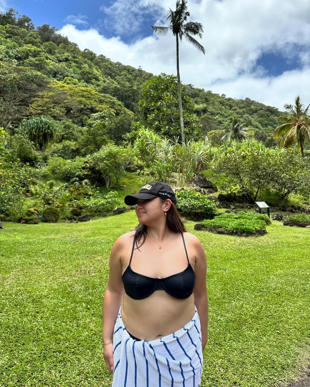 No cenário tropical, uma mulher usa um biquíni simples preto, que destaca sua silhueta de maneira elegante. Ela está envolta em uma toalha com listras brancas e azuis, proporcionando um contraste interessante.