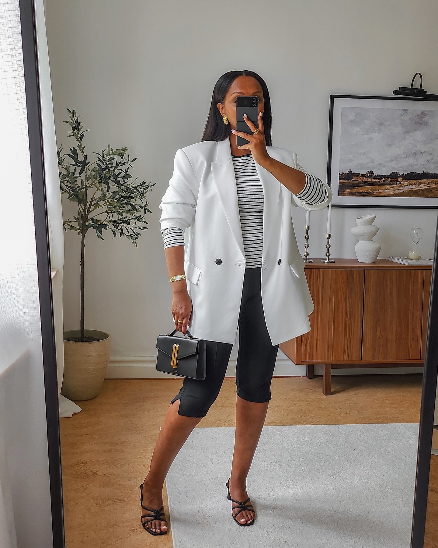 Pessoa vestindo blazer feminino branco, camiseta listrada e calça preta em frente ao espelho, segurando bolsa preta.