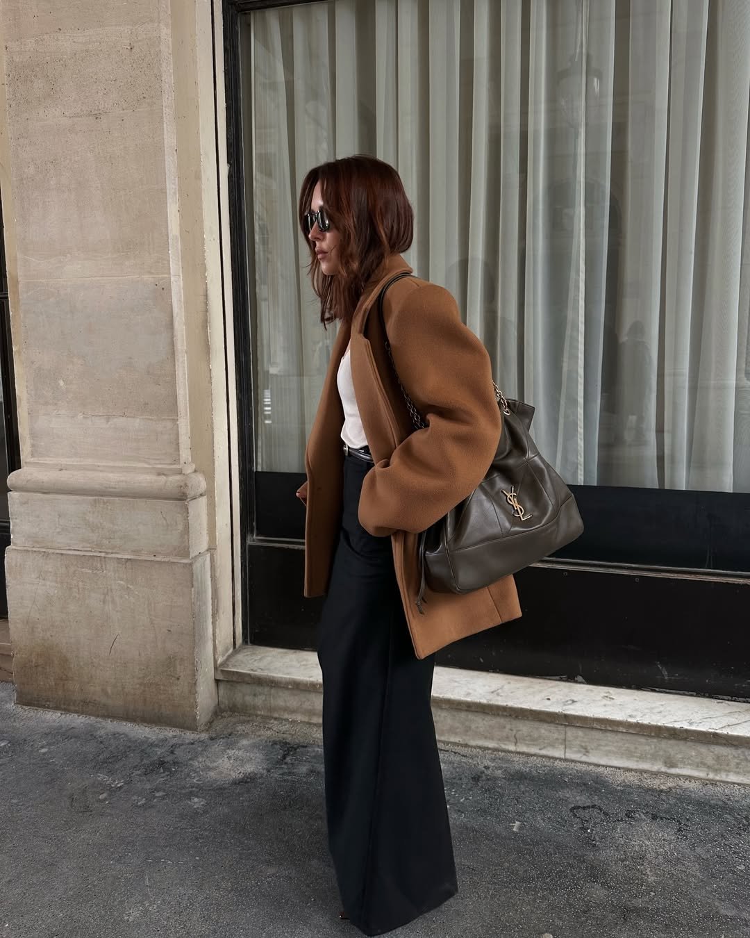 Pessoa usando blazer feminino marrom, calça preta e óculos escuros, carregando bolsa cinza.