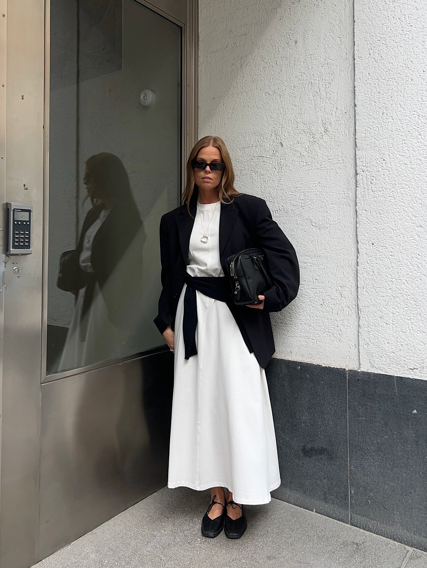 Mulher de vestido branco e blazer feminino preto, usando óculos escuros, encostada em uma parede.