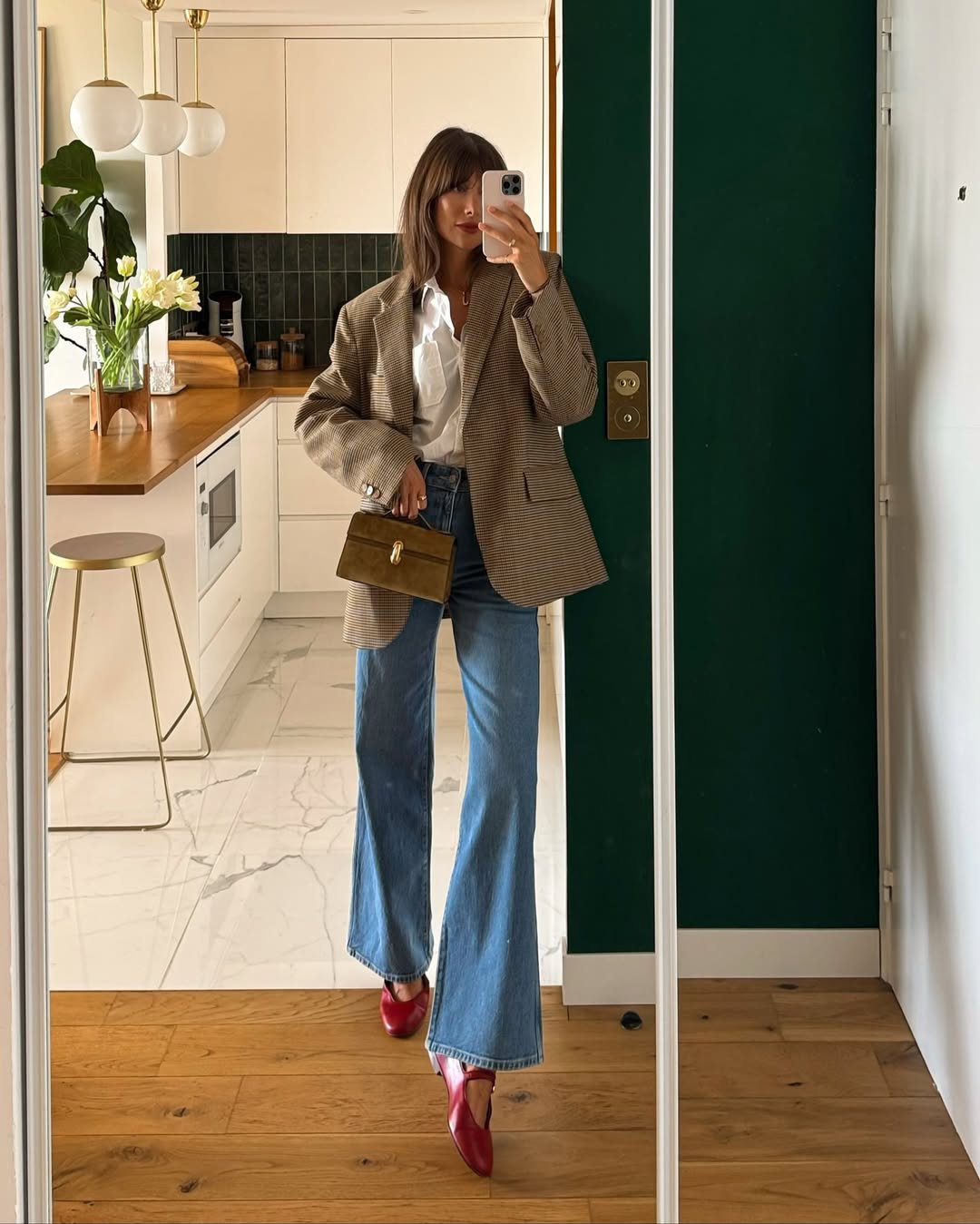 Pessoa usando blazer feminino xadrez, camisa branca, jeans e sapatos vermelhos em uma cozinha moderna.