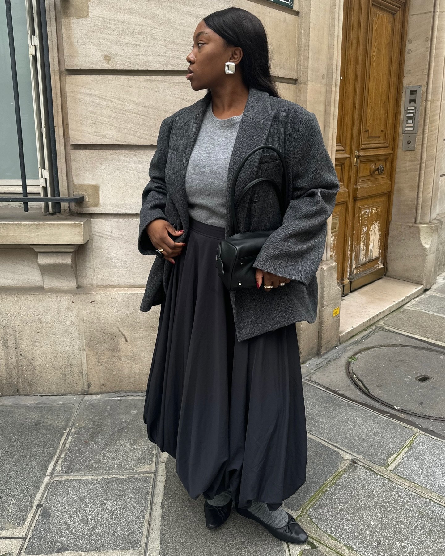 Pessoa com blazer feminino cinza e saia preta, segurando bolsa, posicionada em frente à parede de pedra ao ar livre.