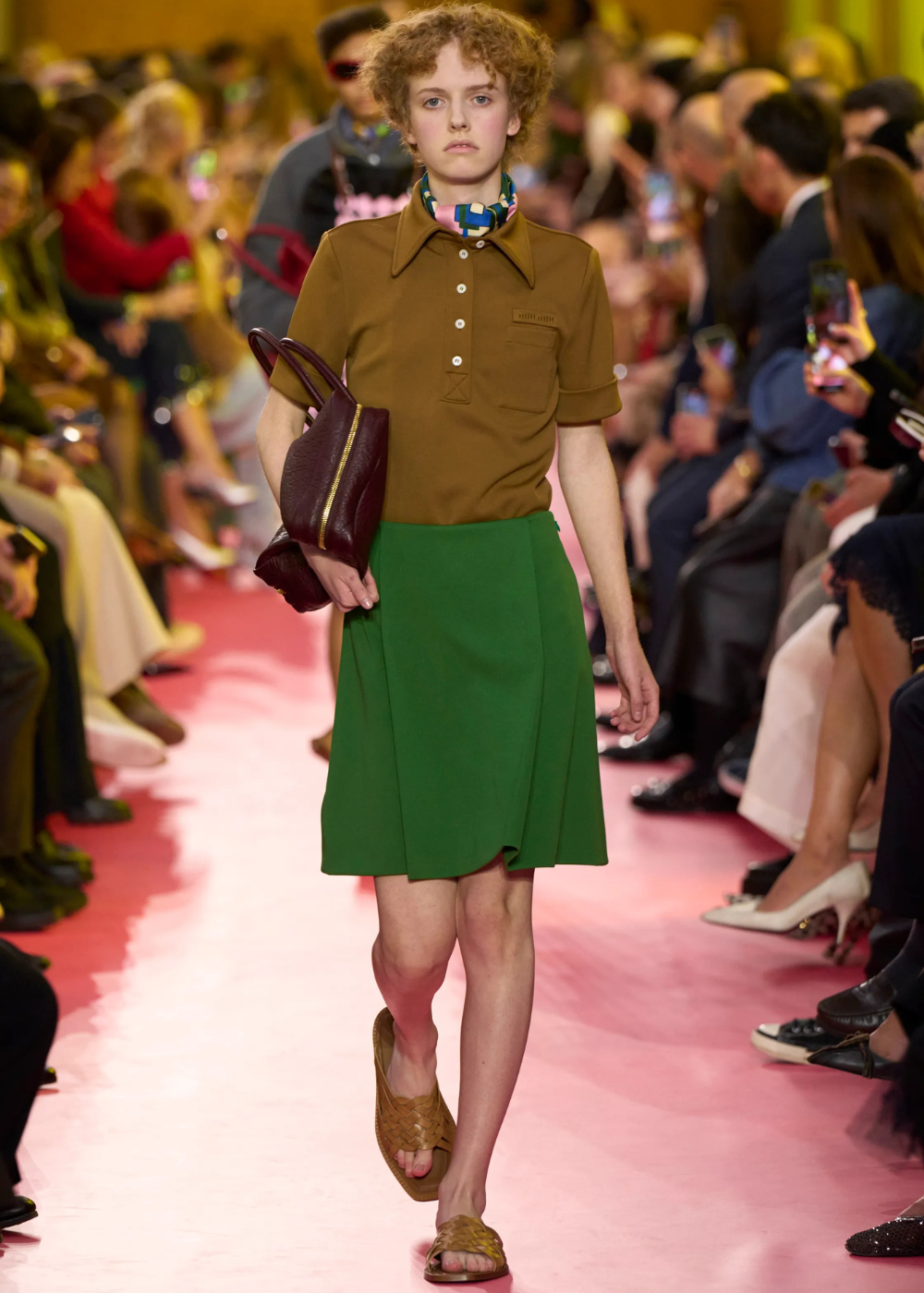 Modelo com blusa polo marrom e saia verde desfila em passarela com bolsa marrom e sandálias.