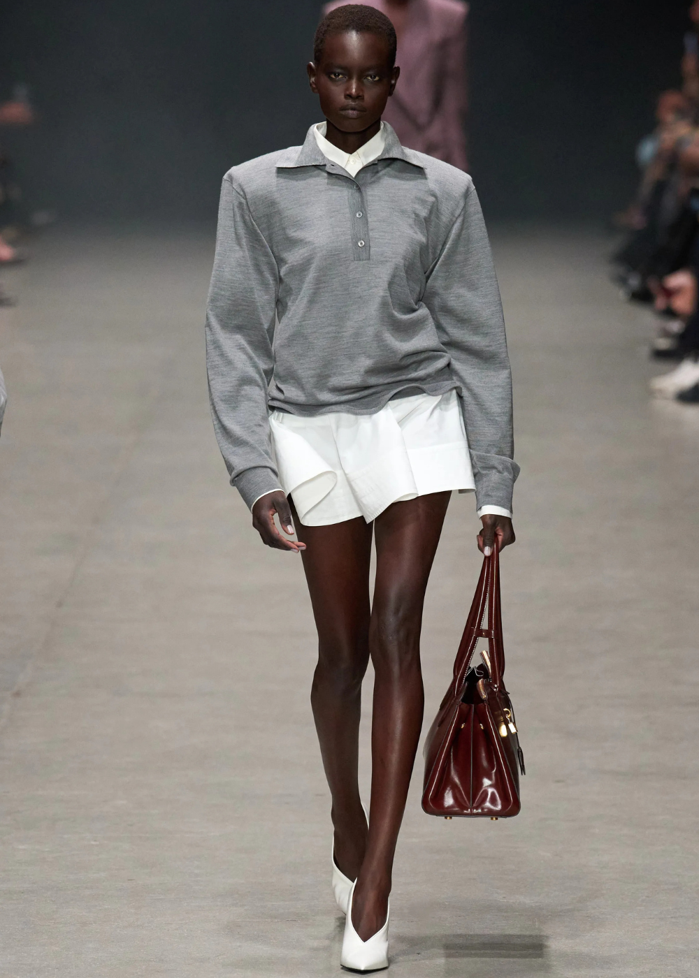 Pessoa veste blusa polo cinza com saia branca e segura bolsa marrom em passarela de moda.