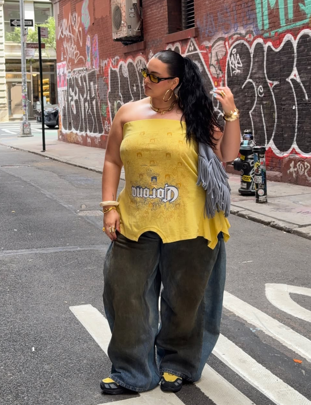 A blusa sem alça em tom amarelo destaca-se por seu design moderno, apresentando estampa gráfica e um corte descontraído. Combinada com calças largas em denim escuro, a peça cria um look ousado e estiloso. 