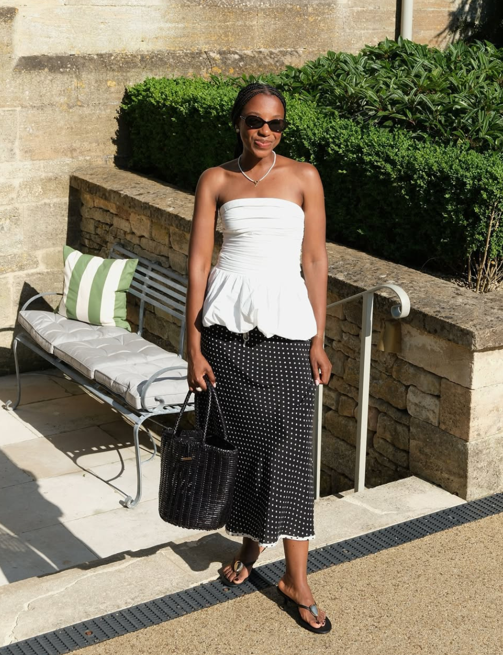 A blusa sem alça em destaque é branca e ajustada ao corpo, com um design peplum que valoriza a silhueta. Combinada a uma saia midi preta com bolinhas brancas, cria um look elegante e descontraído, ideal para dias ensolarados. 