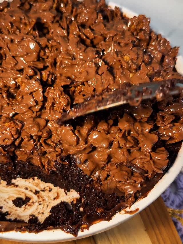 O bolo crocante apresenta uma camada generosa de chocolate derretido sobre uma base macia e úmida. A textura crocante é evidenciada por flocos de chocolate que contrastam com a suavidade do bolo.