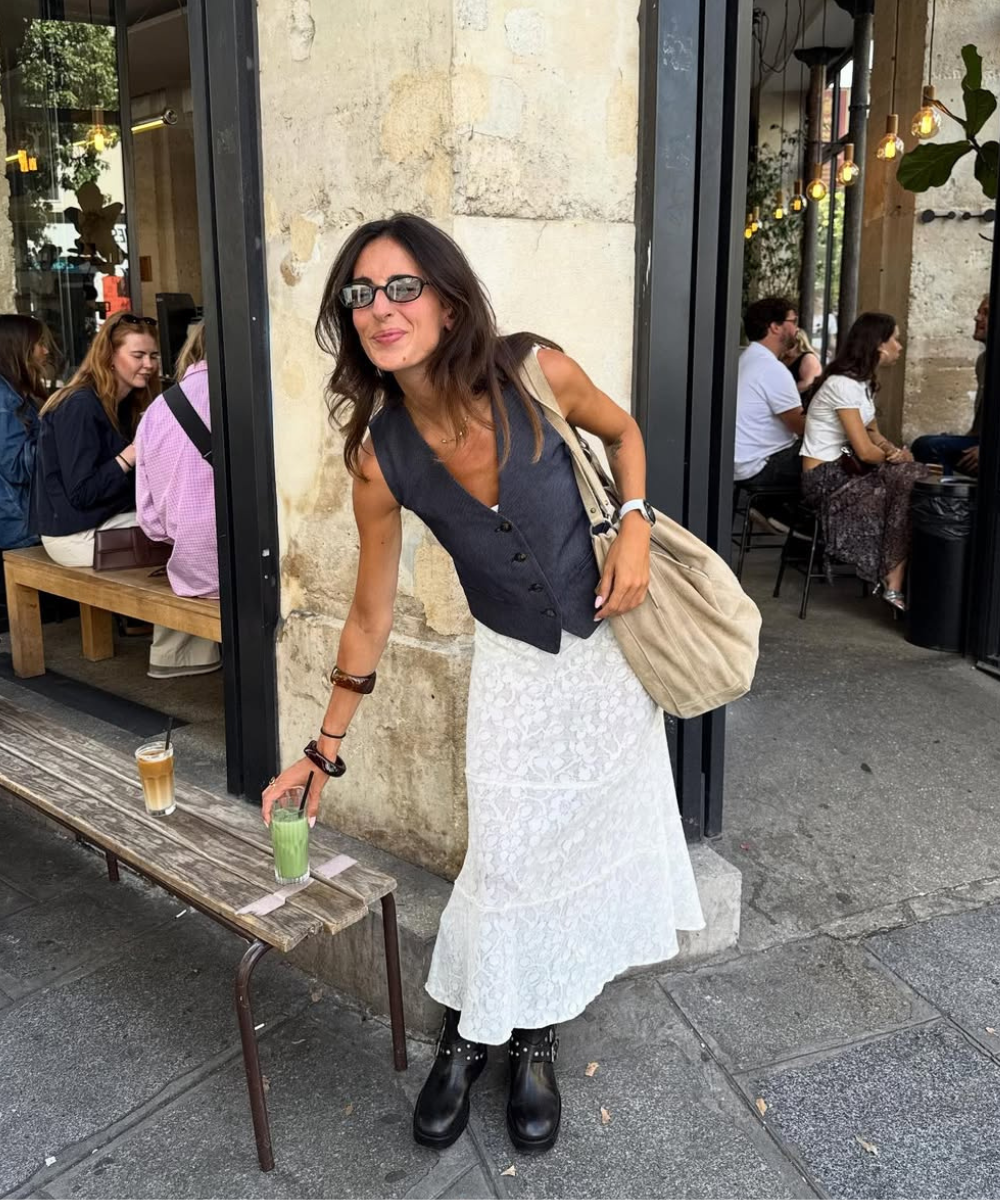 Mulher sorridente com bota, calçado tendência, vestindo colete e saia branca floral, segura um copo de bebida verde em frente a um café.