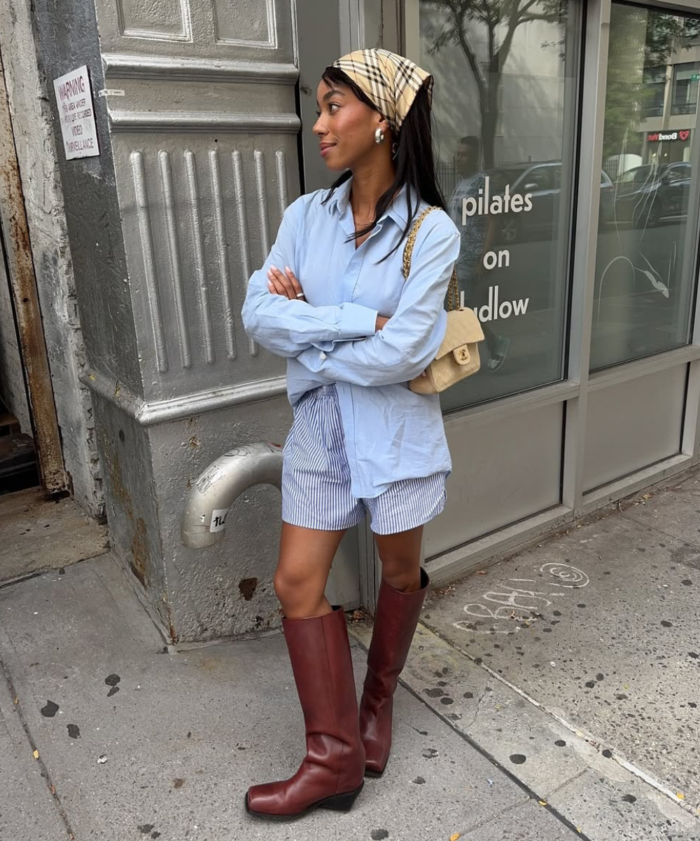 Jovem de lenço na cabeça e botas marrons modernas em frente a edifício urbano. Calçado em destaque como tendência.