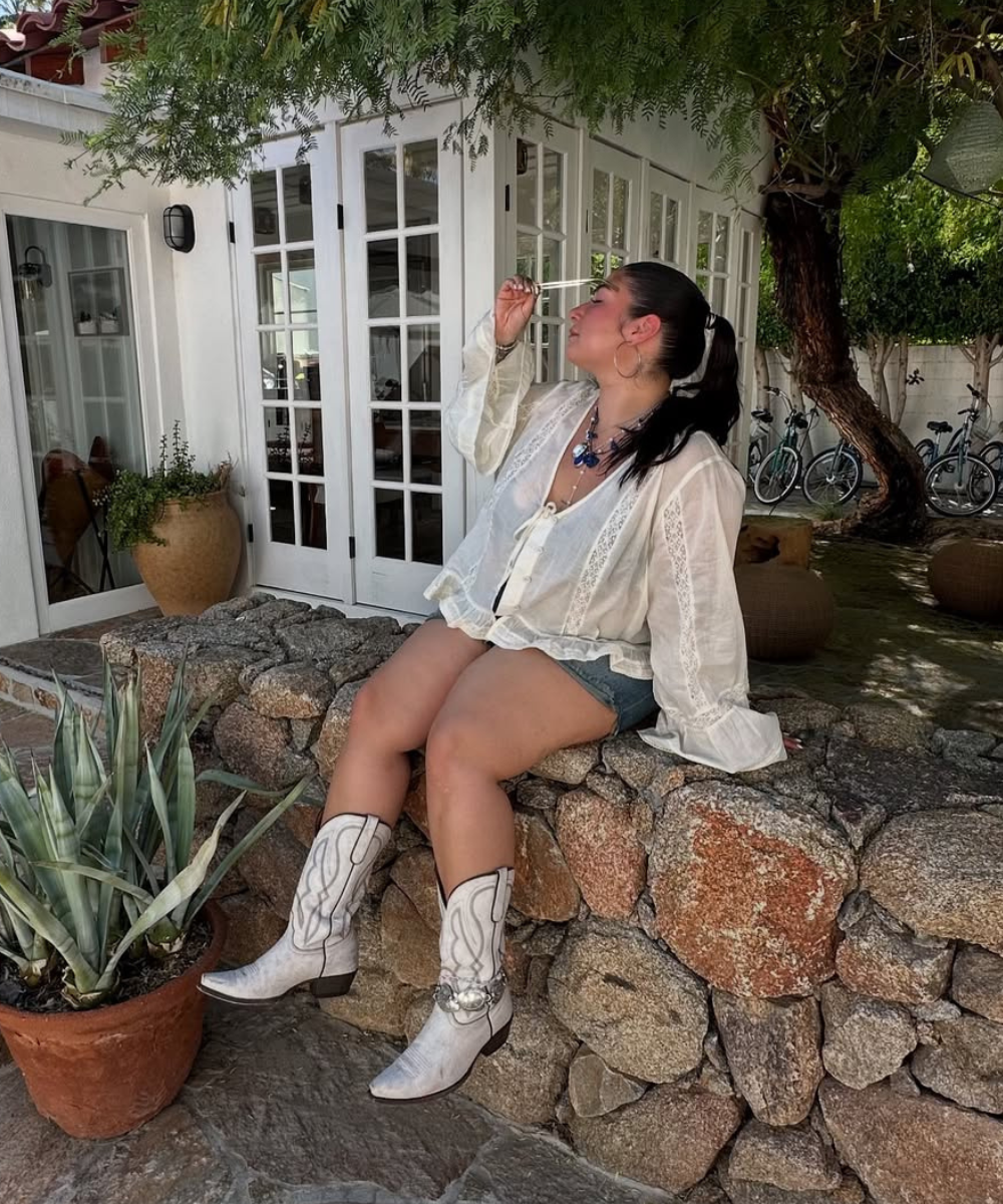 Mulher sentada em muro de pedras, usando botas brancas, calçado tendência, e roupa leve em ambiente externo.